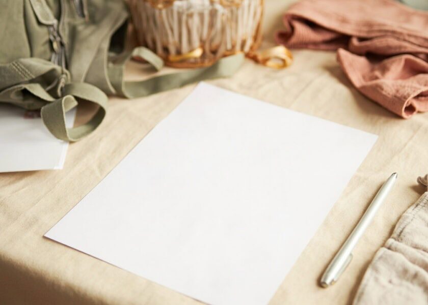 A blank white sheet rests on a cream table beside a silver pen, a small backpack, a basket and folded clothes.