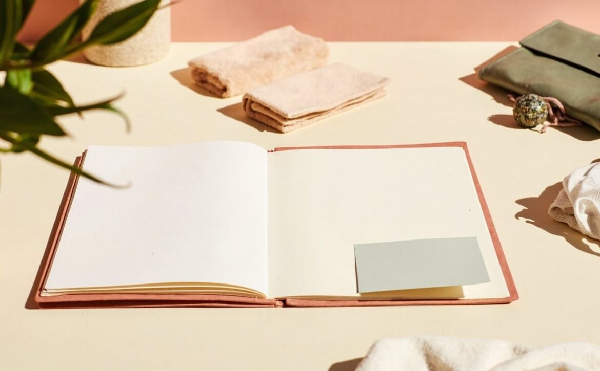 Warm still life of an open blank folder with a plain card, folded textiles, a closed wallet and soft greenery.