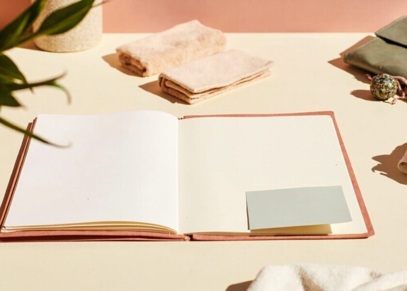 Warm still life of an open blank folder with a plain card, folded textiles, a closed wallet and soft greenery.