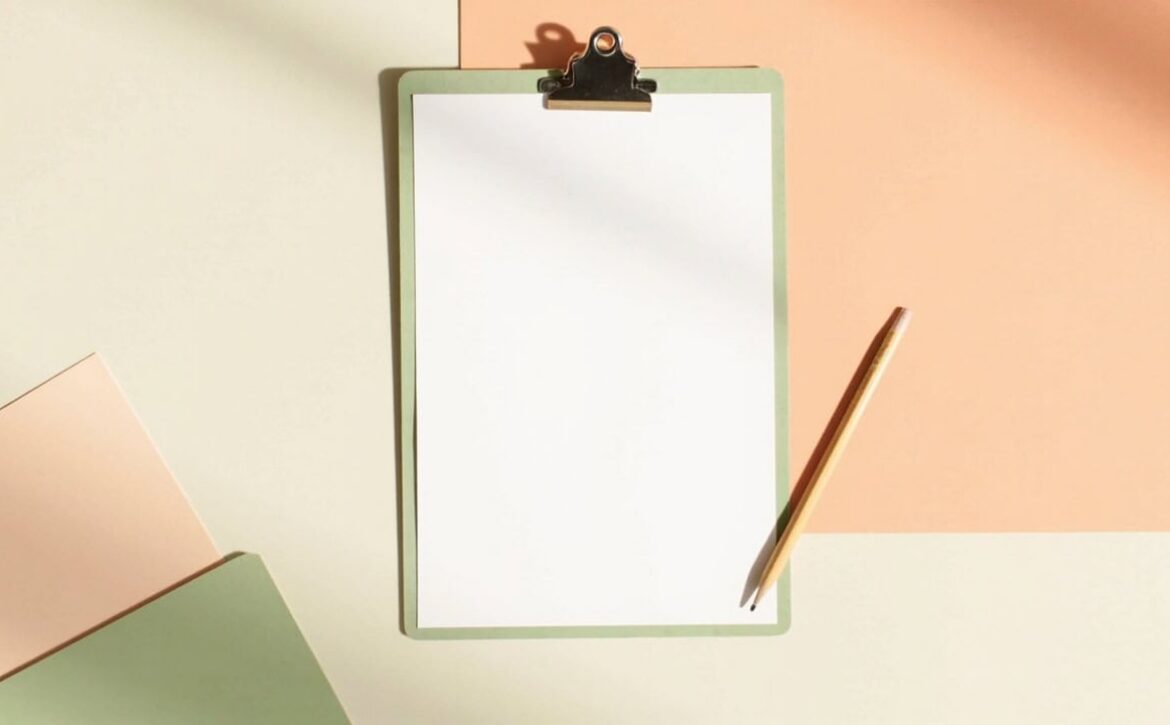 A blank clipboard, an unmarked pencil and layered sage and peach folders on a warm cream tabletop in soft light.