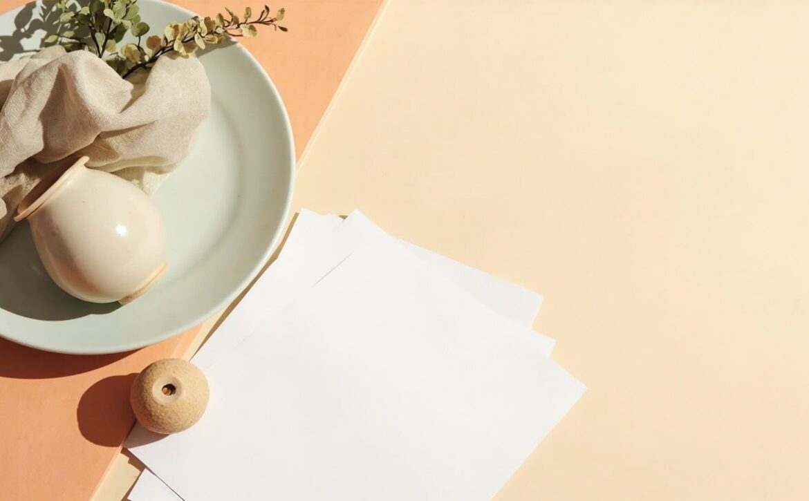 Blank papers, folded fabric, a rounded ceramic vessel, greenery and a wooden bead in warm natural light.