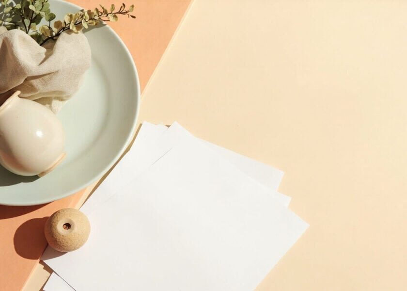 Blank papers, folded fabric, a rounded ceramic vessel, greenery and a wooden bead in warm natural light.