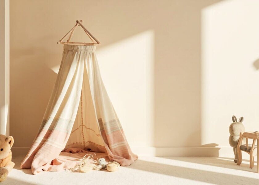 A cream fabric play canopy in a sunlit neutral room, beside a teddy, scattered toys, a plush rabbit and miniature wooden furniture.