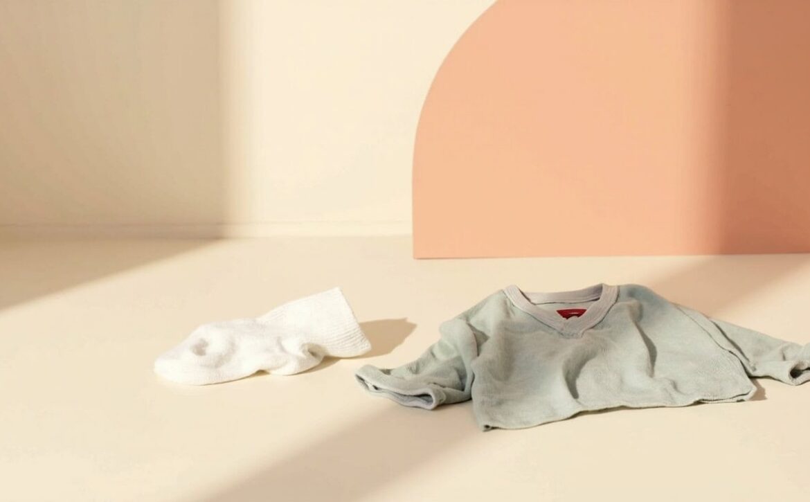 A pale sage child's shirt and white sock rest on a cream surface against a soft coral backdrop.