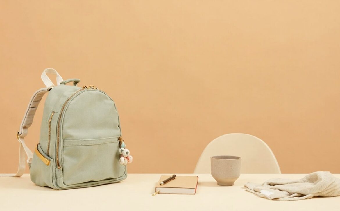 A sage school satchel with an unmarked notebook, ceramic cup and folded garment on a cream table in warm beige tones.