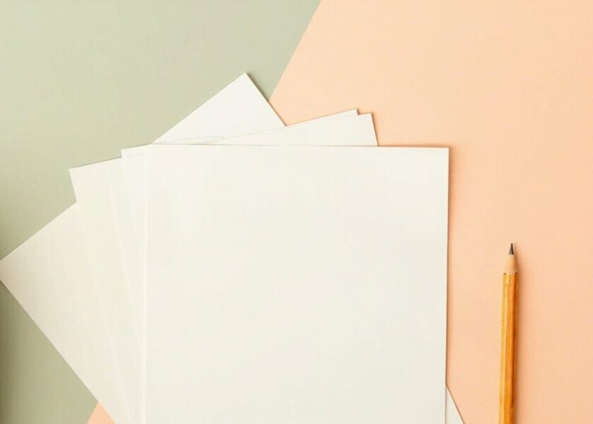 Blank sheets fanned beside a yellow pencil and a cropped, unbranded beige satchel on a sage-and-coral background.