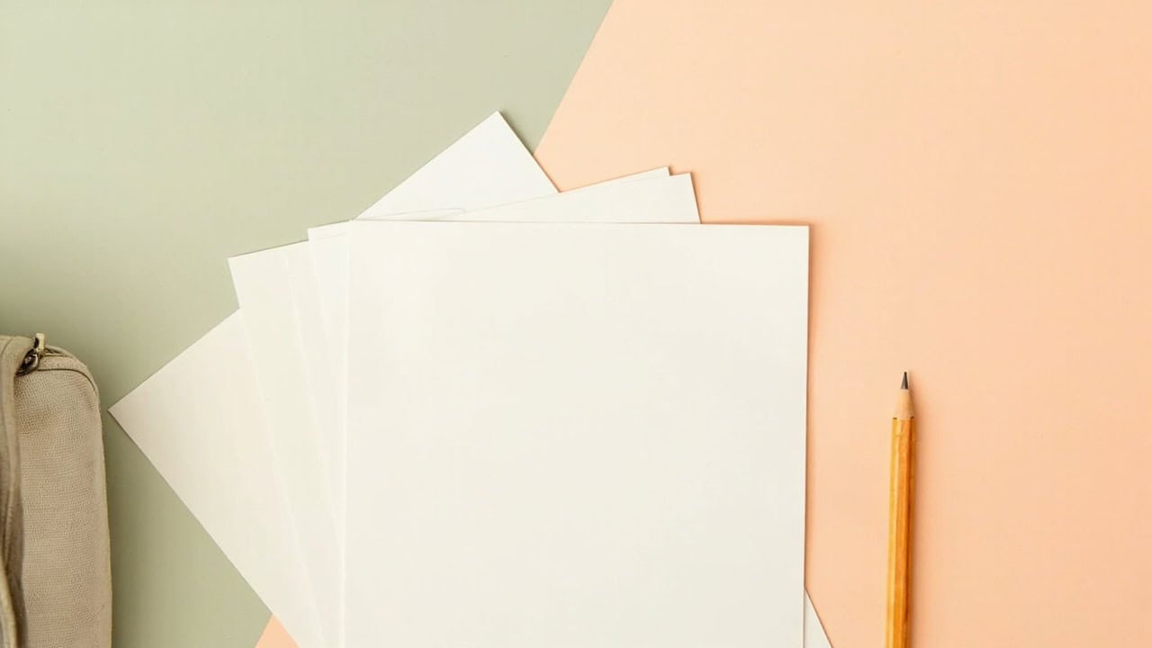 Blank sheets fanned beside a yellow pencil and a cropped, unbranded beige satchel on a sage-and-coral background.