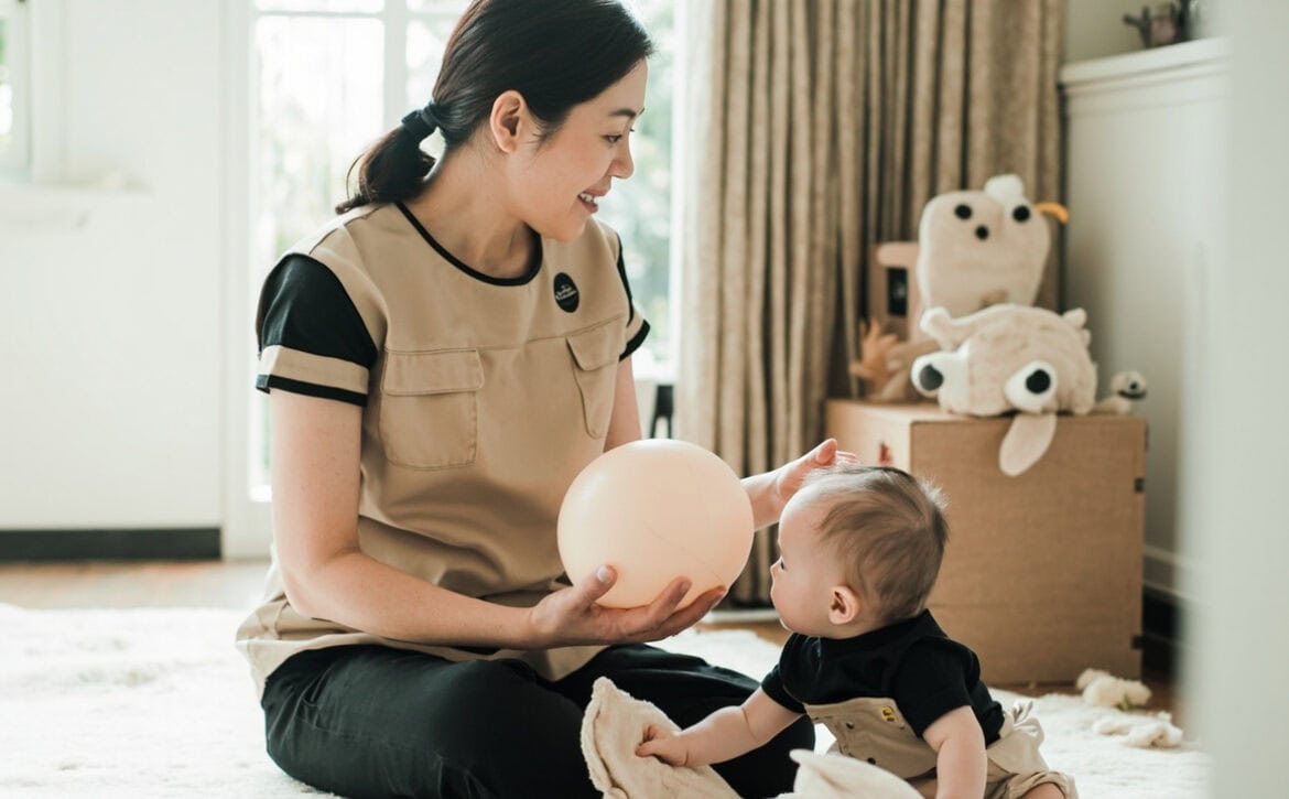 A seated caregiver in a beige sleeveless tunic over a black top presents a soft ball beside an awake baby seated on a floor mat.