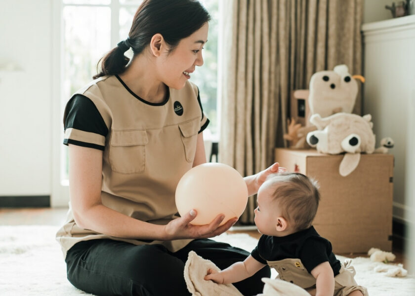 A seated caregiver in a beige sleeveless tunic over a black top presents a soft ball beside an awake baby seated on a floor mat.