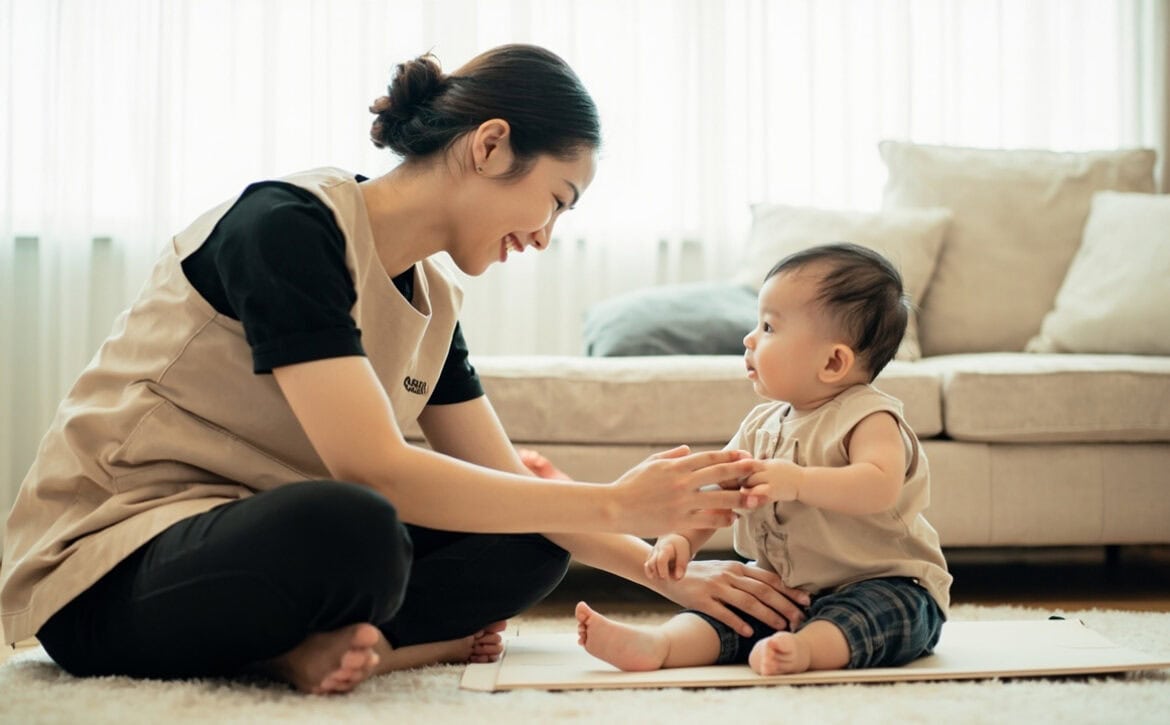 A caregiver in a beige tunic sits cross-legged beside an awake baby, supporting the baby's seated posture while holding one hand.