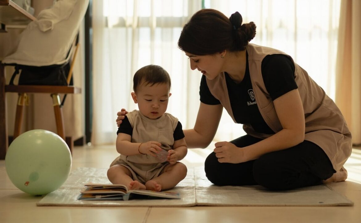 A uniformed caregiver kneels beside a seated baby on a floor mat, with an open board book and a large soft ball nearby.