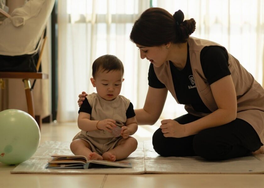 A uniformed caregiver kneels beside a seated baby on a floor mat, with an open board book and a large soft ball nearby.