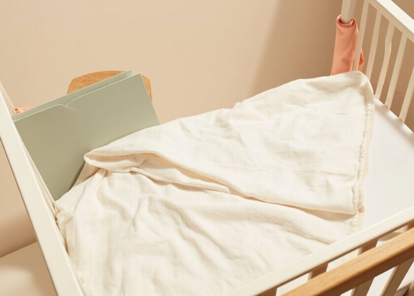 A warm cream cot with a soft muslin blanket, closed sage folder and small coral fabric accents.