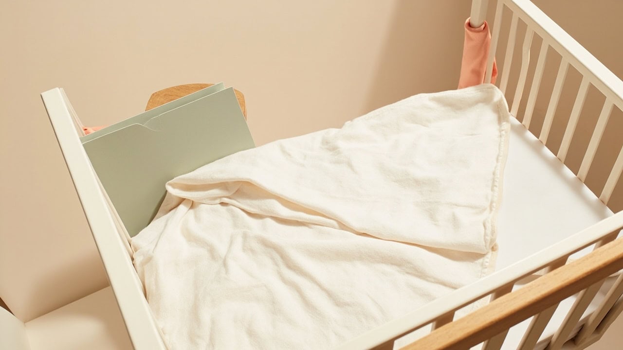A warm cream cot with a soft muslin blanket, closed sage folder and small coral fabric accents.