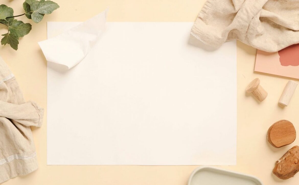 Blank paper, wooden play pieces, neutral fabric, greenery and a partial ceramic dish arranged on a warm cream surface.