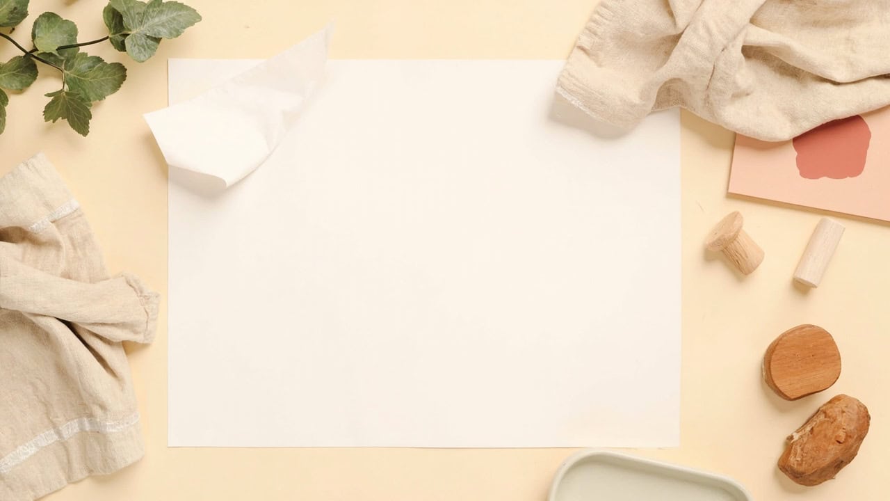 Blank paper, wooden play pieces, neutral fabric, greenery and a partial ceramic dish arranged on a warm cream surface.