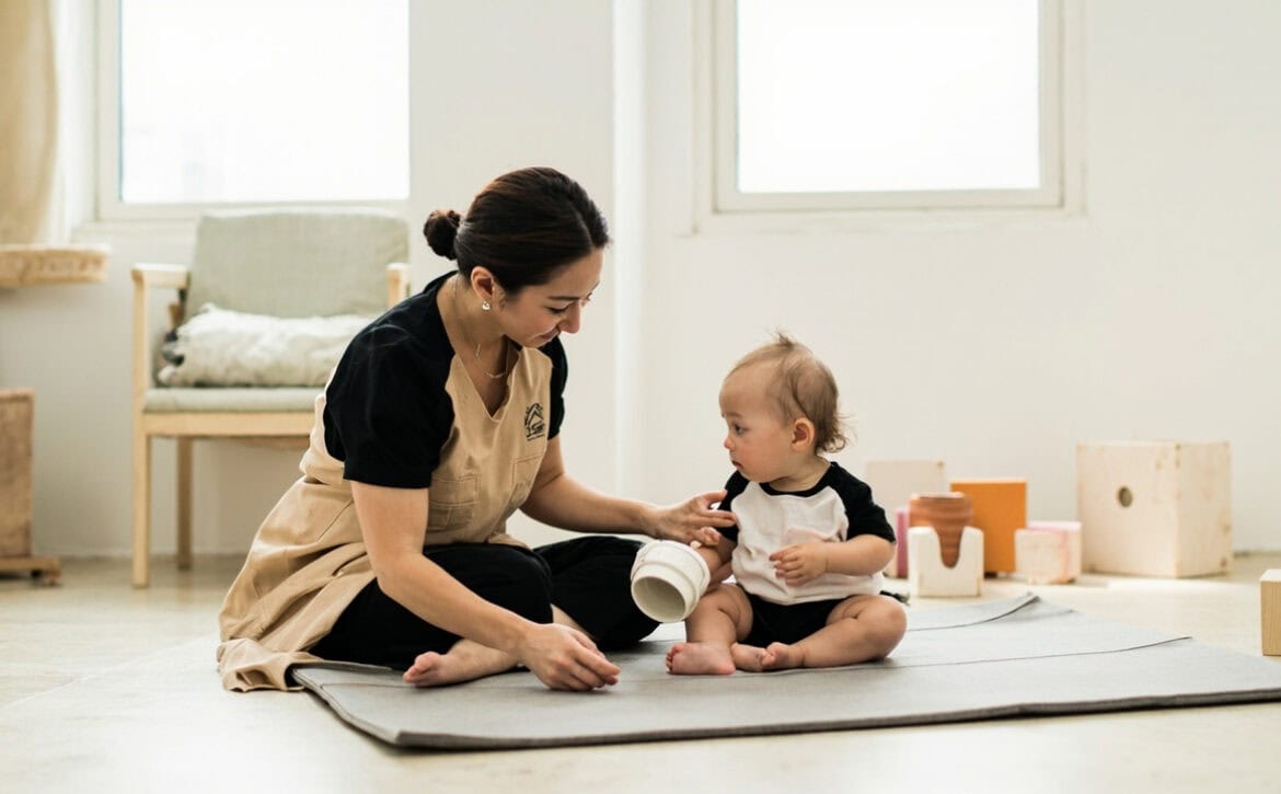 A caregiver kneels beside a seated toddler on a play mat, offering a white cup while the toddler holds a pink cup in a bright room.