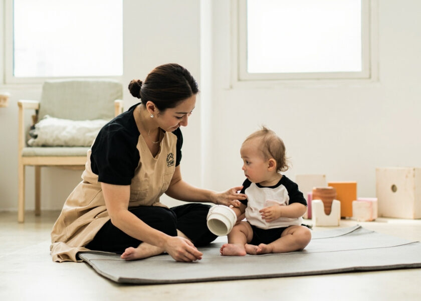 A caregiver kneels beside a seated toddler on a play mat, offering a white cup while the toddler holds a pink cup in a bright room.