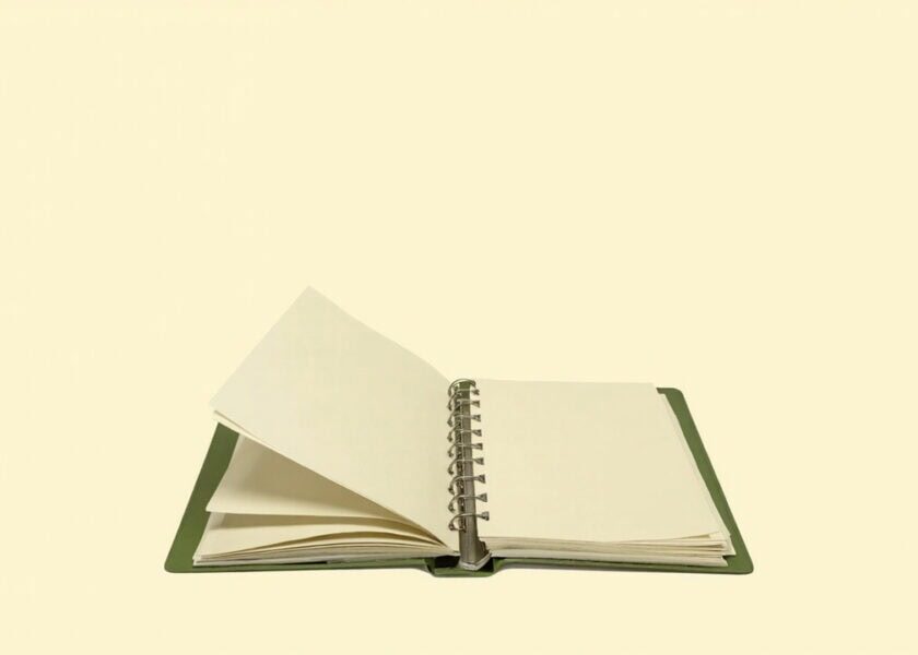 An open muted-green ring binder with blank cream pages sits alone on a warm ivory background.