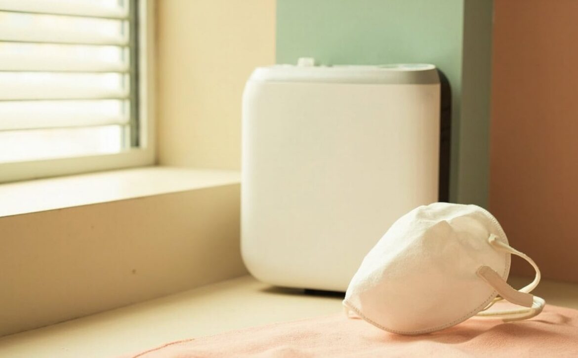 An unmarked white cup-shaped respirator rests on a coral cloth beside a louvred window and a smooth, generic air purifier.