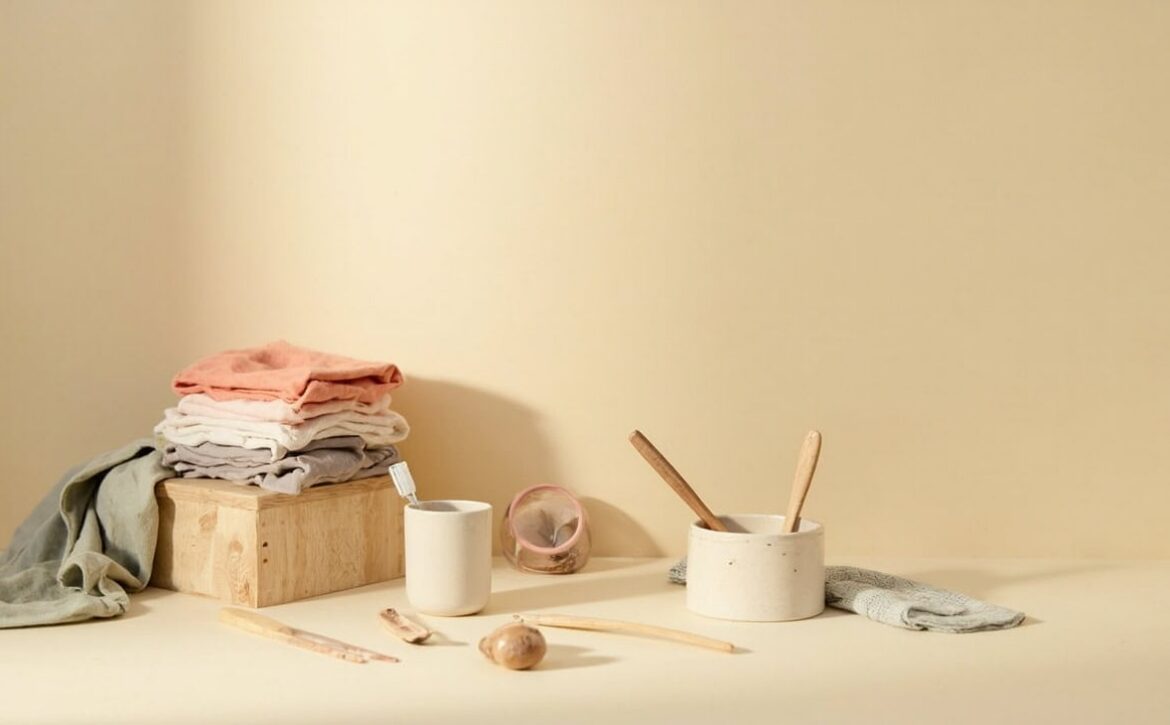 Warm cream still life of folded clothes, a toothbrush in a cup, ceramic bowl, wooden utensils and cloths.