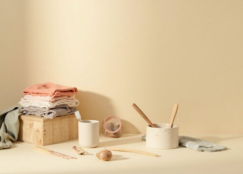 Warm cream still life of folded clothes, a toothbrush in a cup, ceramic bowl, wooden utensils and cloths.