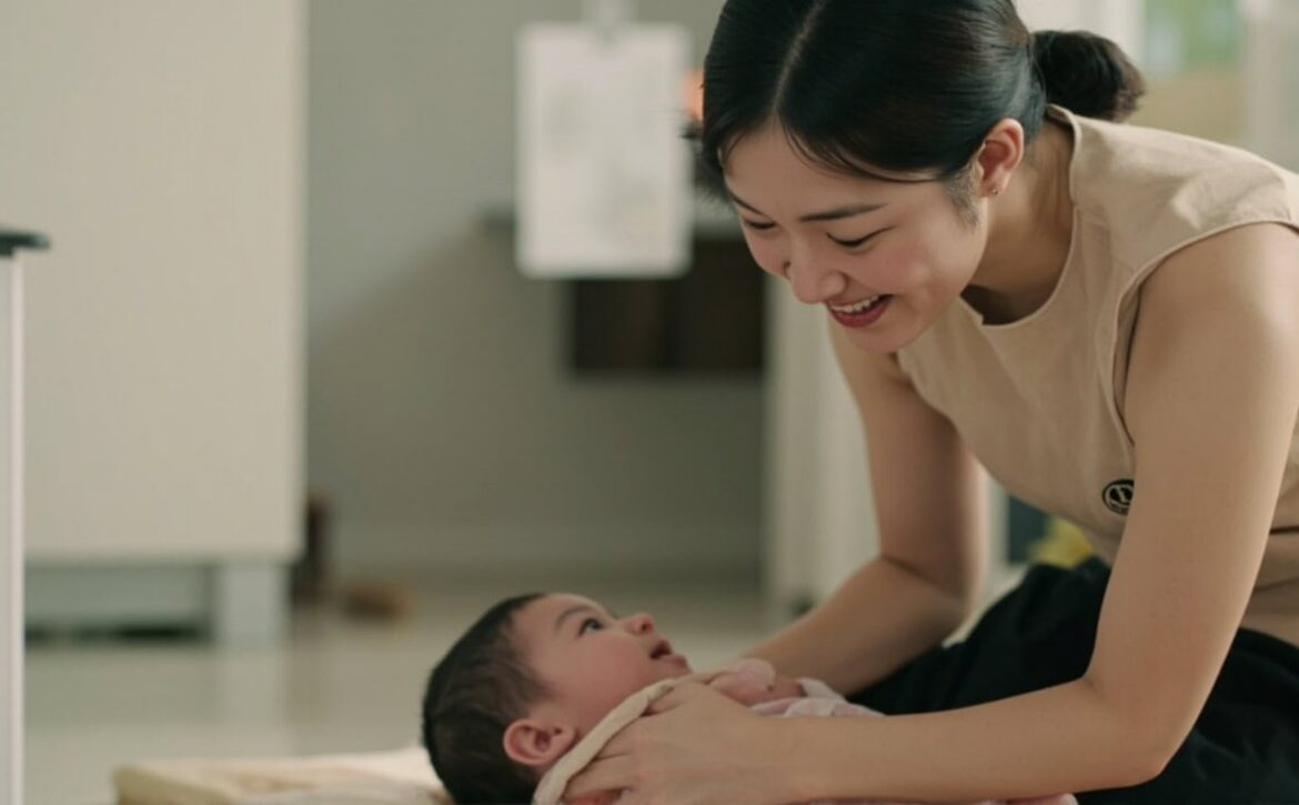 A smiling adult leans towards an awake baby lying on a mat and looking up at them.
